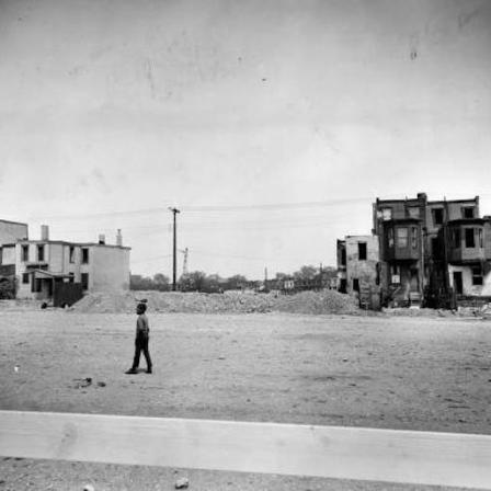 black and white photo. child walking in the foreground. row homes are on the right and left in the background with a pile of rubble and bulldozer in between them.